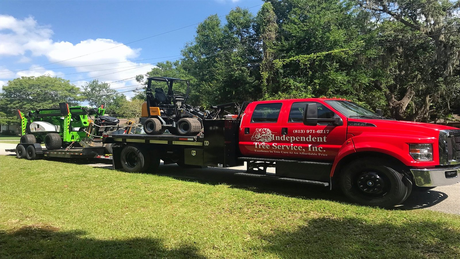A red truck from Independent Tree Service Inc. transports tree-cutting equipment on a sunny day, surrounded by lush greenery and trees.