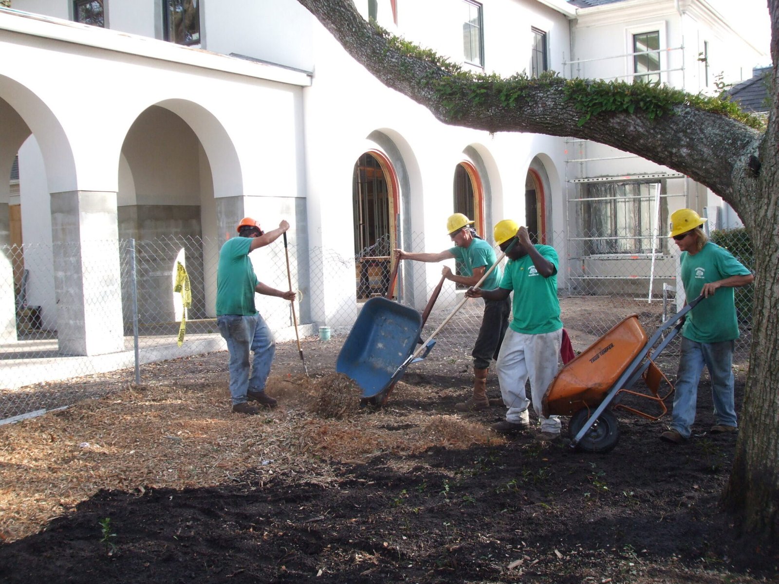 Four people in green shirts and yellow helmets work with wheelbarrows and shovels on a mulch-covered area near an arched building.